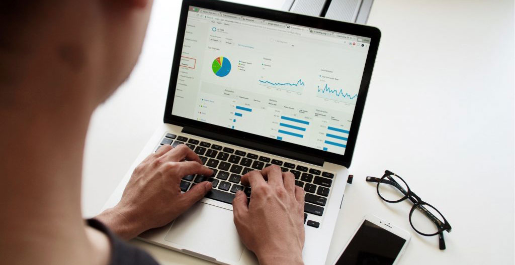 Des mains tapent sur un clavier d'ordinateur portable à côté d'un carnet de notes ouvert avec des post-its colorés sur le bord de l'écran