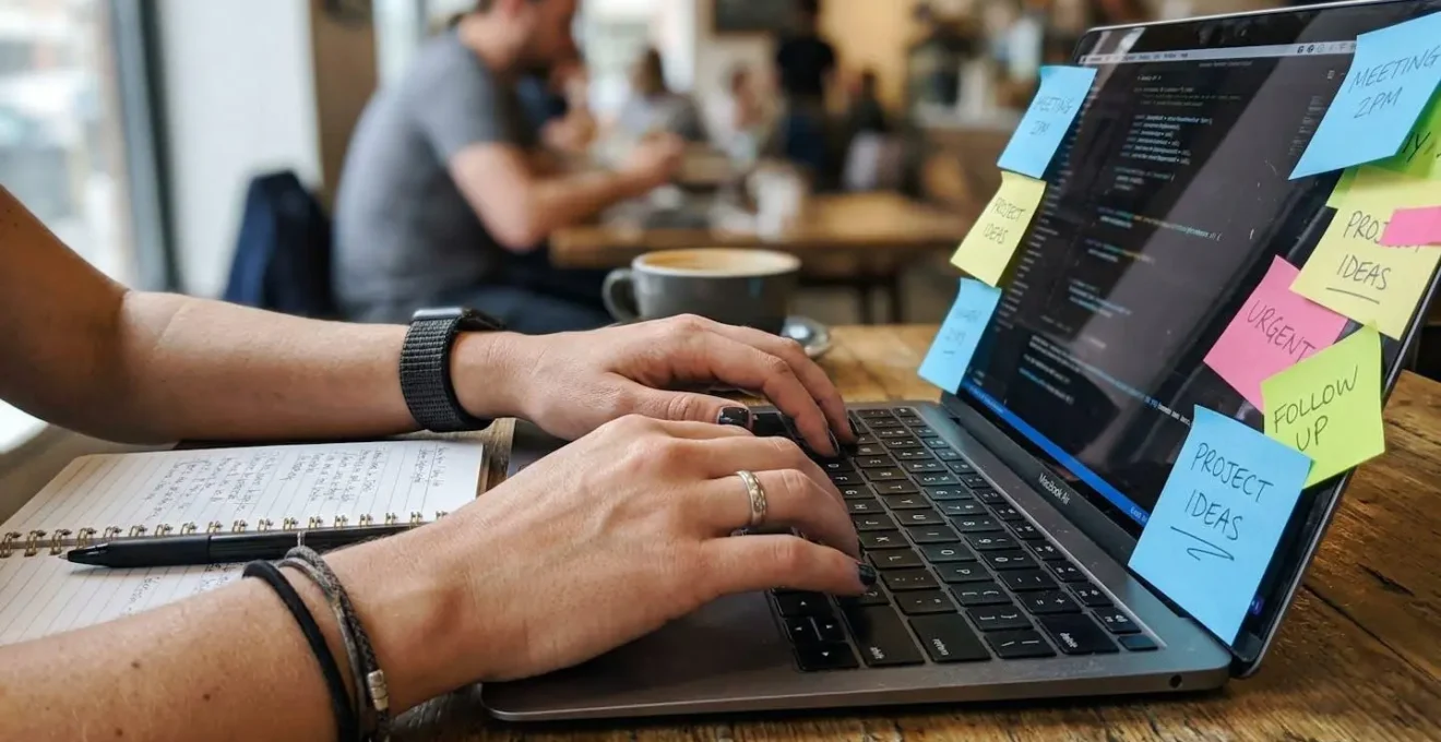 Des mains tapent sur un clavier d'ordinateur portable à côté d'un carnet de notes ouvert avec des post-its colorés sur le bord de l'écran